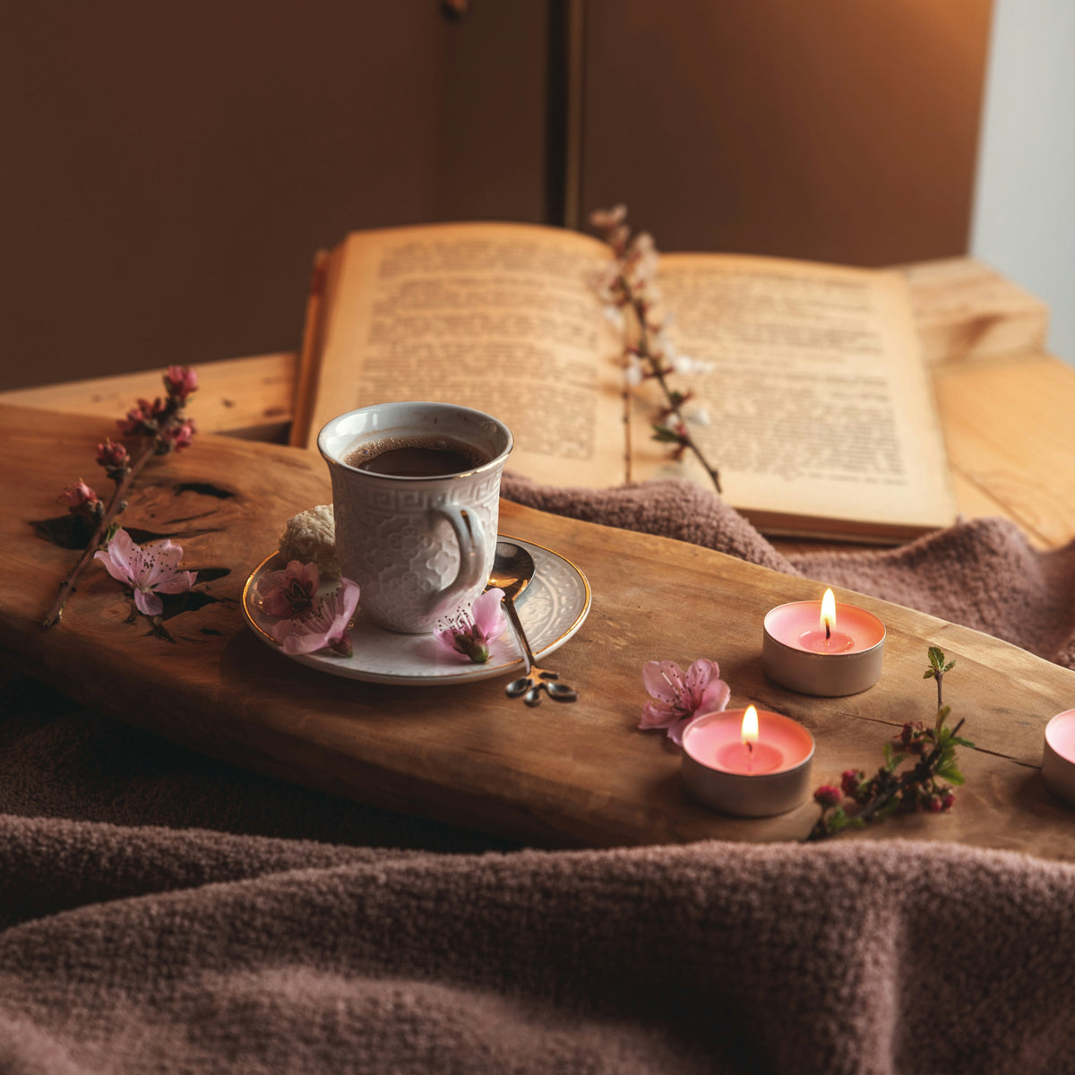 Cup of herbal tea with saucer, spoon, and candles on a wooden surface with an open book in the background.